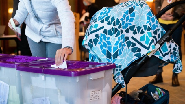 Woman's arm putting a piece of paper in a voting box, next to a pram covered in a patterned blue shawl