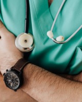 Image of a man's arm with a watch on, the man is wearing green medical scrubs and has a stethoscope around his neck